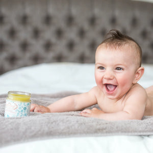 Anointment Baby Balm on bed beside smiling baby
