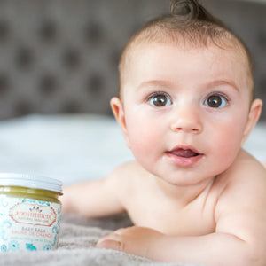Anointment Baby Balm on bed beside baby with brown eyes